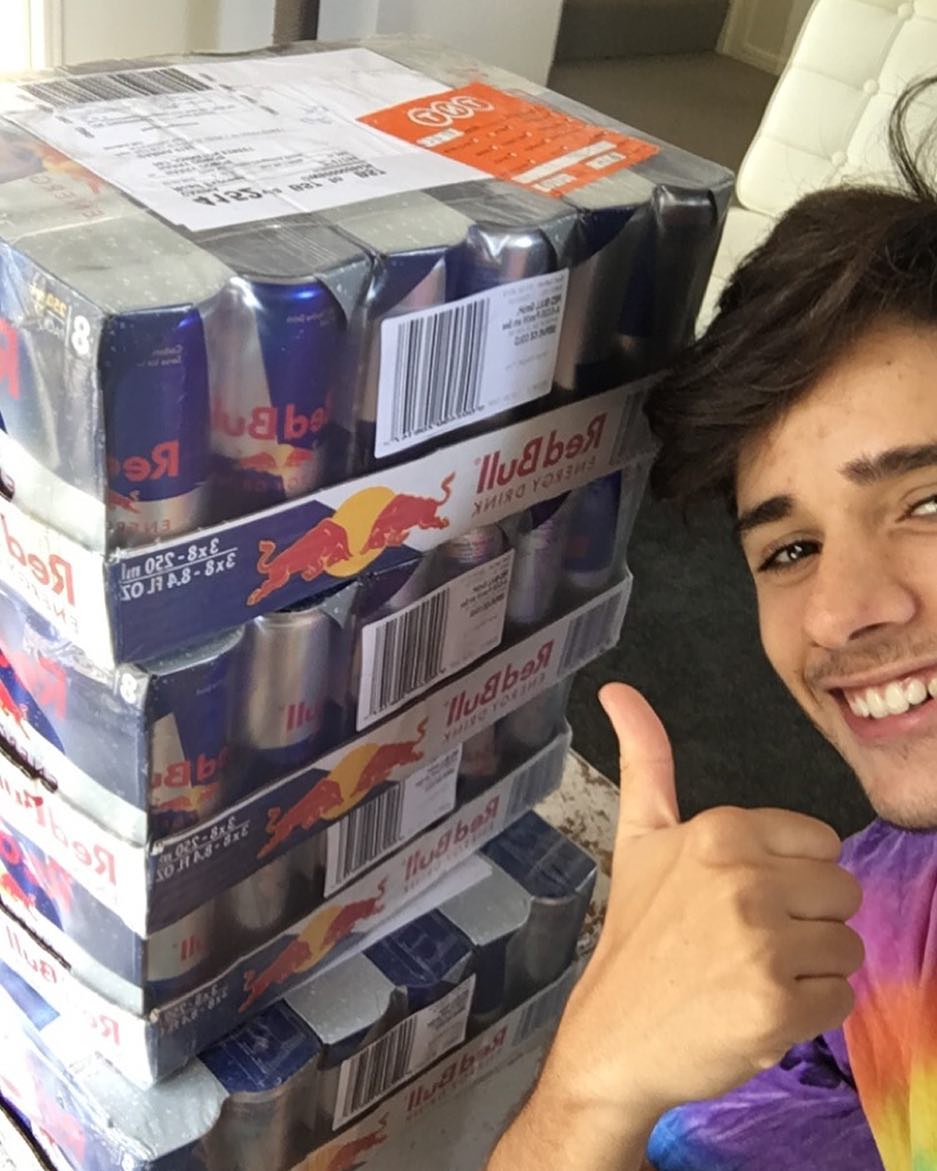 Harry Hope grinning with a thumbs up next to a shoulder-high stack of Red Bull slabs