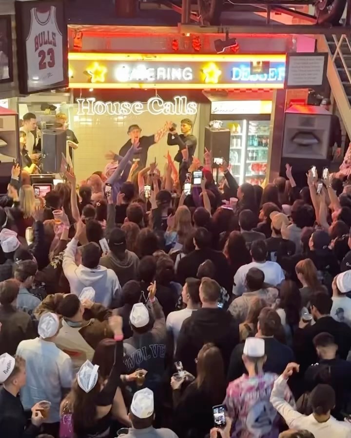 A packed crowd in paper deli hats facing a tiny stage under a House Calls neon sign